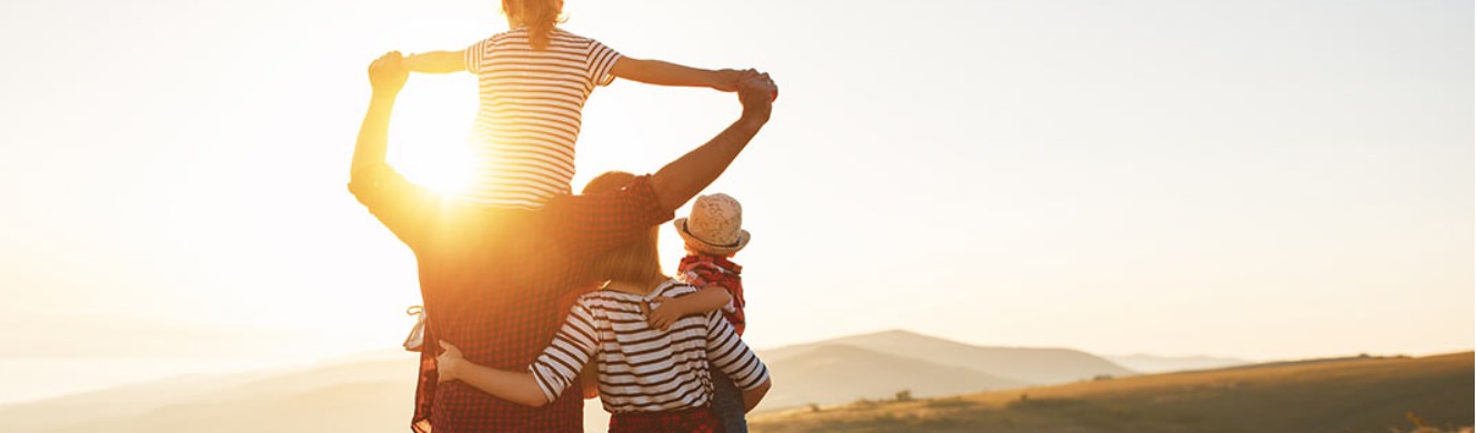 Dad with girl on his shoulders and mom holding baby looking at a scenic sunset 